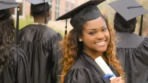 Image of a college graduate in a black cap and gown holding a diploma facing the camera while other graduates face the other direction