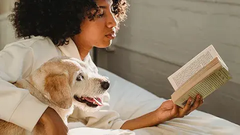 A woman reads a book on a bed with her arm around a dog.