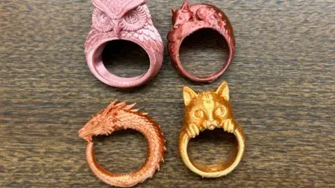 Four 3D printed rings: a pink owl, a burgundy fox, an orange dragon, and a golden cat resting on a counter.