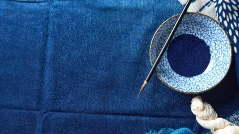 Fabric in various shades of blue topped with a patterned bowl containing blue powder and paint brush and blue and white fabric along the top and bottom.