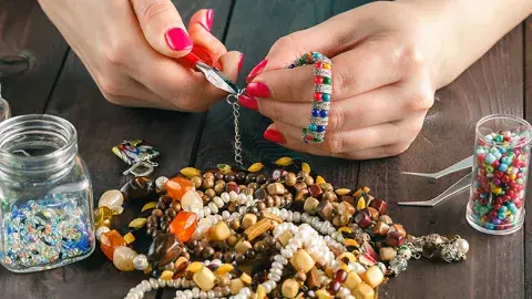 A pair of hands makes a piece of jewelry with pliers and assorted beads.
