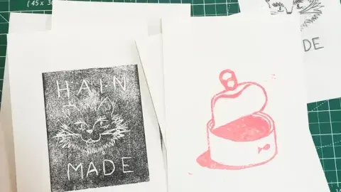 Example linocut prints on cards, one with a cat face that says "Hain Made" and another with an open can of food.