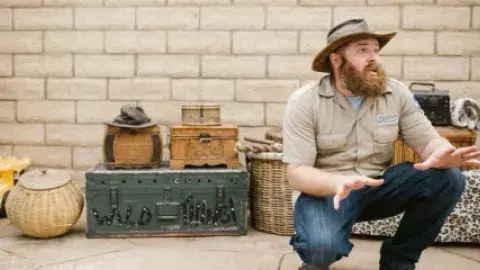 A bearded adult man crouched in front of a block wall with various baskets and trunks behind him.