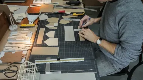 Person measuring and cutting cardboard pieces on a gridded work table with various crafting tools and materials around.