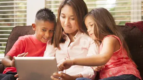 a mother holds a computer tablet in her lap, sharing it with a young boy and girl sitting on either side