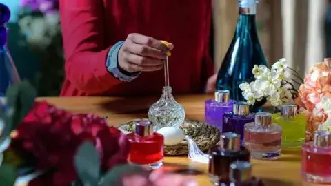 Person at table with perfume making supplies and various perfume bottles and vessels.