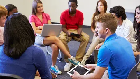 Group of teens sitting in a circle holding laptops and iPads.