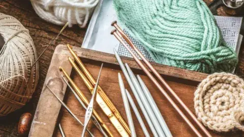 Spools of different coloredd yarn and twine sit on a wooden table. A wooden tray holds metal tools for knitting and crocheting.