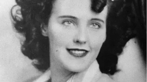 Black and white photo of Elizabeth Short, better known as the Black Dahlia, smiling and gazing past camera wearing a light colored suit jacket.