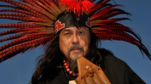 Performer Martin Espino holds an instrument and wears a feathered red headdress.