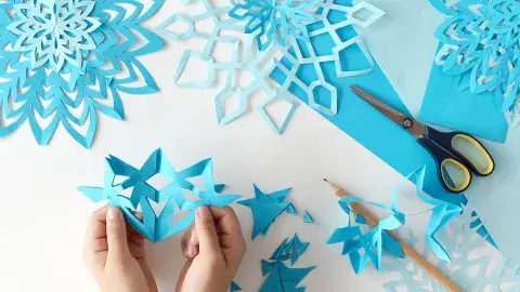 Hands hold a paper cut snowflake with blue snowflakes, scissors and paper on a table.