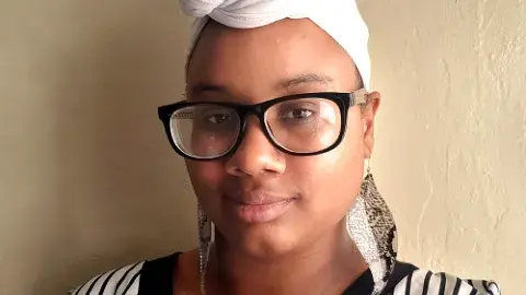 Diamond Forde wearing glasses and a head wrap, leaning against a wall