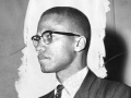 Black and white photo of Malcolm X from the side, standing at a meeting.