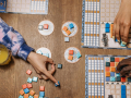 Image of 3 hands reaching from various points across a table with a board game set up. Turquoise and orange tiles are organized on different parts of the table and game board.