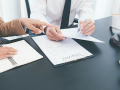 The hand of a person in a suit points to a document held by another person, next to a gavel and scales
