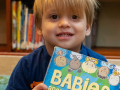 Toddler seated on colorful rug in children's section of library, holding a board book with illustration of babies, titled "Babies."