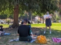 librarian reads a picturebook to a group of seated toddlers and adult caregivers in a park with trees