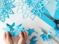 Two hands holding blue paper shaped like a snowflake, various hues of blue craft paper in the background, scissors and a pencil also in the background.
