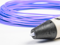 A partial view of a white 3D pen with a black tip angled at the bottom right corner and a spool of purple plasic filament behind it on a white surface.