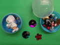 Picture of two capsule containers, one open and one sealed, with various beads and other colorful items in them.