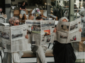Three ladies sitting in chairs in a row holding up newspapers in front of their faces