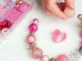 Hands assembling a pink beaded bracelet beside a tray of beads and jewelry supplies.