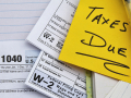 a yellow post it note with Taxes Due and three tax forms on top of a notebook