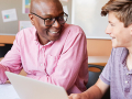 An adult mentor smiles while helping a teen student work on a laptop at a table with notebooks.