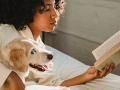 A woman reads a book on a bed with her arm around a dog