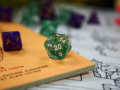 A green 20-sided die sits on a table with other dice and game pieces behind it. 