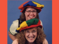 Two women standing together with colorful hats and stage costumes.