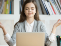 Person sitting in front of a laptop with bookshelves in the background. The person's hands are raised with thumb and forefingers touching and their eyes are closed.