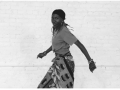 black and white photo of a Black woman standing in a dance pose wearing a boldly printed cloth skirt with a solid t-shirt
