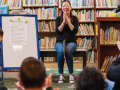 A woman sitsin front of a group of toddlers and adults clapping her hands. A white board is next to her facing the crowd. 