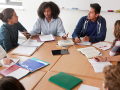 Group of multi racial teens, in discussing, sitting around classroom desks with folders and notebooks open. 
