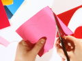 In the foreground a person's hands are holding scissors and pink craft paper with a heart drawn on it. The person is cutting out the heart. The background shows other cut-out hearts of different colors. 