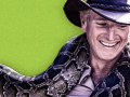 A smiling man in a safari hat holds a boa constrictor around his neck