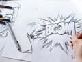 A person’s hand is shown drawing the word boom in bold letters with a pencil on a blank sheet of white paper