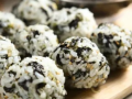 A group of rice balls mixed with seaweed and sesame seeds are arranged on a wooden board with a fork in the background.