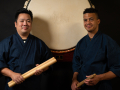 Two BIPOC men in blue wrap coats, holding drumsticks, standing in front of a large taiko drumhead