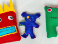 Three hand sewn dolls made of felt against a white background. The doll on the right is a red square with big spiked yellow hair, black button eyes, and a square mouth with yellow lips and light blue zipper The middle doll is dark blue, skinny, and bear-shaped with one button eye on the left and a stitched red X on the right. The doll on the right is a green square with two legs, short pink spikes on it's head, two pink button eyes, and a black uneven line for its mouth