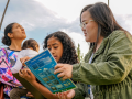 In an outdoor area with palm trees, a female adult with straight dark hair, eyeglasses and a green jacket is holding an instruction guide on clouds with a young girl with curly brown hair. Another female with lght brown curly hair in bun and holding a toddler over her shoulder is to the left of them looking at a blue sky with partial cloud cover and the sun above.
