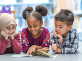 Three diverse children lay on their bellies on the floor, looking at a shared book