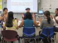 A group of adults sitting in a circle in chairs, drumming a rhythm with their hands