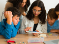 Five teens are gathered around a circuit board and looking at it with interest. They are surrounded by plyers and other tools.