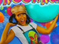 Cheri Jacobs of Snooknuk and Friends wearing a white t-shirt and colorful alien print overalls with a multi-colored balloon backdrop. 