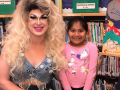 A smiling drag queen in a blonde wig poses in the library with two smiling children
