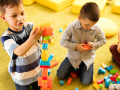 Three young children are playing with colorful building blocks on a soft, yellow carpet.
