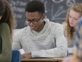 Two female students and two male students are sitting at desks in a high school classroom taking a test with a chalkboard with writing in the back.