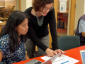 woman with black long sleeve tutor top and blue plants working with a learner wearing blue and white pattern shirt. The tutor is standing and point to the lesson instructing the learner during a lesson.
